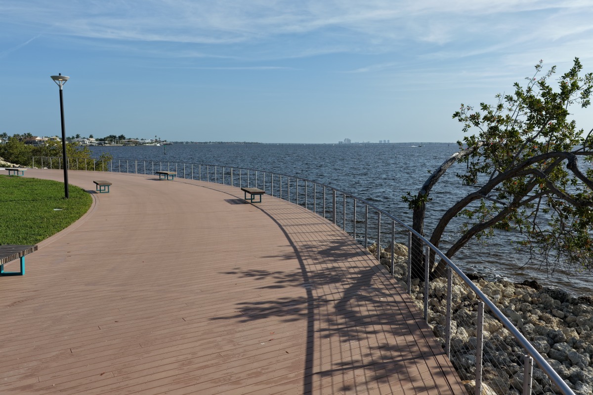 Curving wooden boardwalk winds through newly planted palms and greenery with benches along the path.